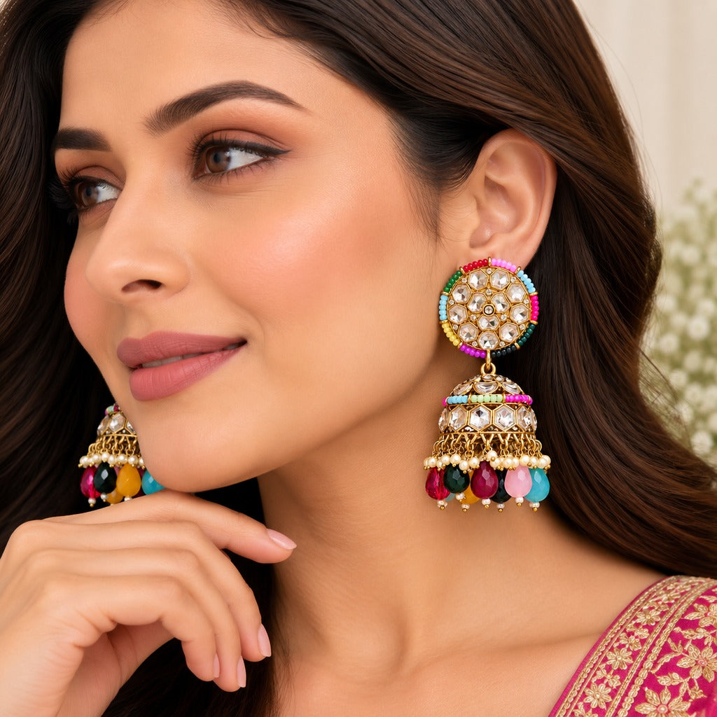 A close-up lifestyle shot of a woman wearing ornate gold-plated Indian jhumka earrings with colorful gemstone accents and dangling bell-shaped charms.